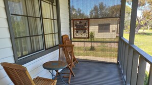 Une terrasse en bois avec deux chaises et une petite table, une fenêtre avec des rideaux et une vue sur un espace verdoyant.