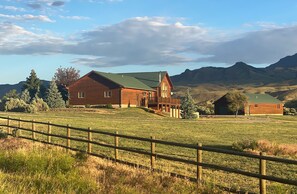 Un grand bâtiment en bois, surmonté d’un toit végétalisé, entouré d’un champ clôturé et avec des montagnes en arrière-plan.