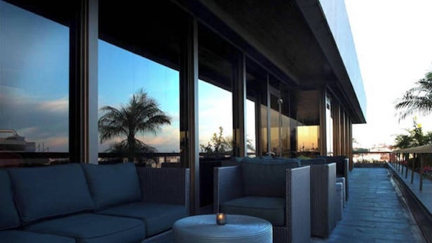 Un área de estar exterior moderna con sofás y mesas redondas, grandes ventanales que ofrecen vistas a palmeras y un atardecer.