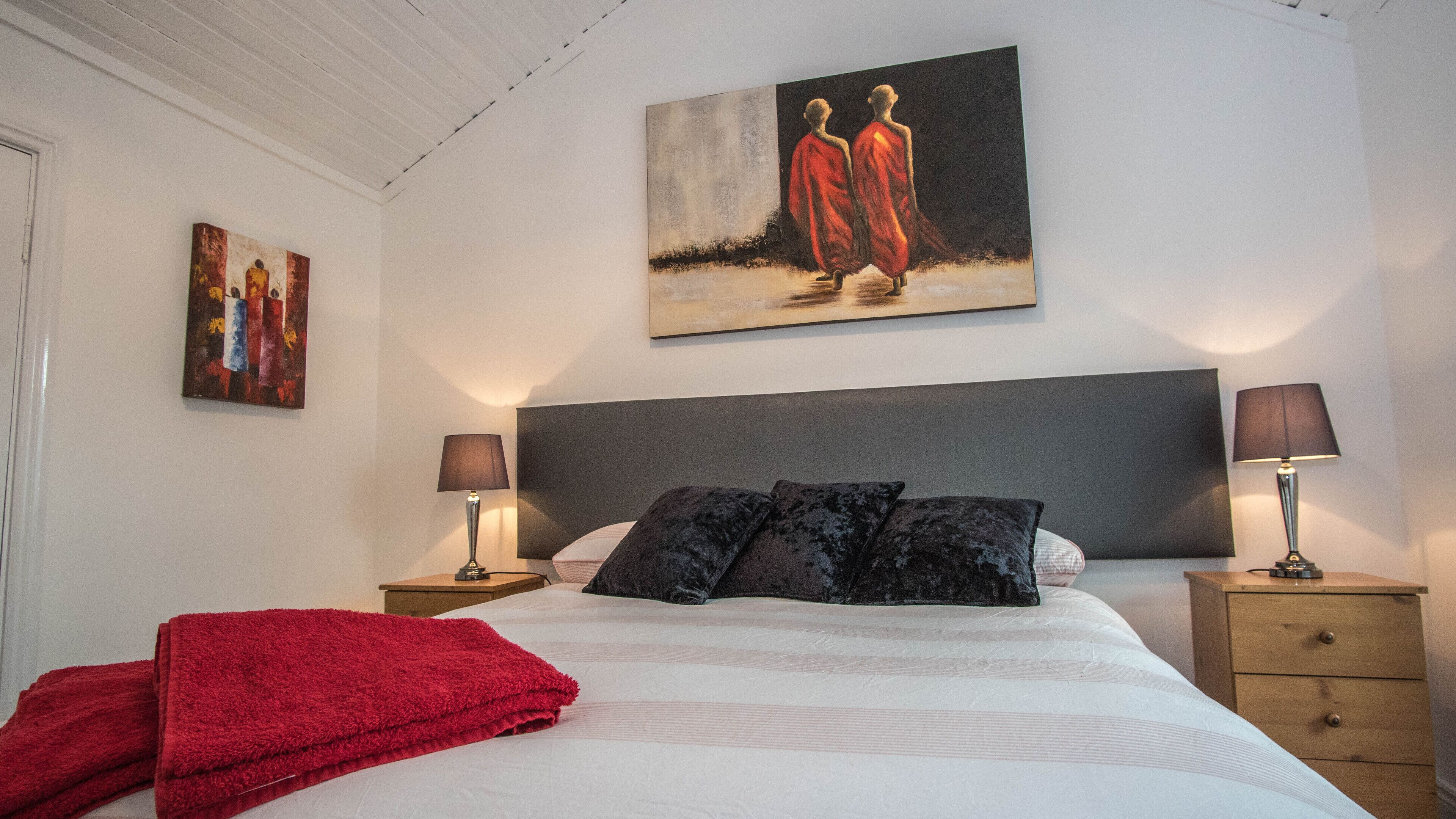A bedroom with a large bed, two bedside tables, a red throw blanket, and artwork on the walls.