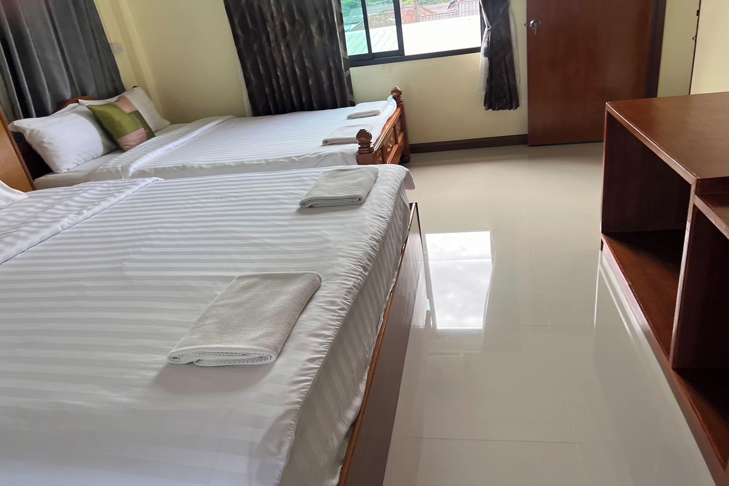 Habitación de hotel con dos camas, cada una con una toalla blanca doblada con cuidado. La habitación tiene un cabecero de madera, una mesita de noche de madera y un ventanal con cortinas.
