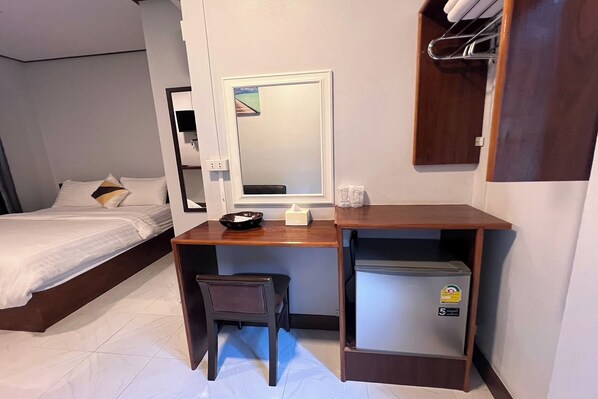 A hotel room with a bed, a desk with a chair, a small refrigerator, a mirror, and a shelf with towels.