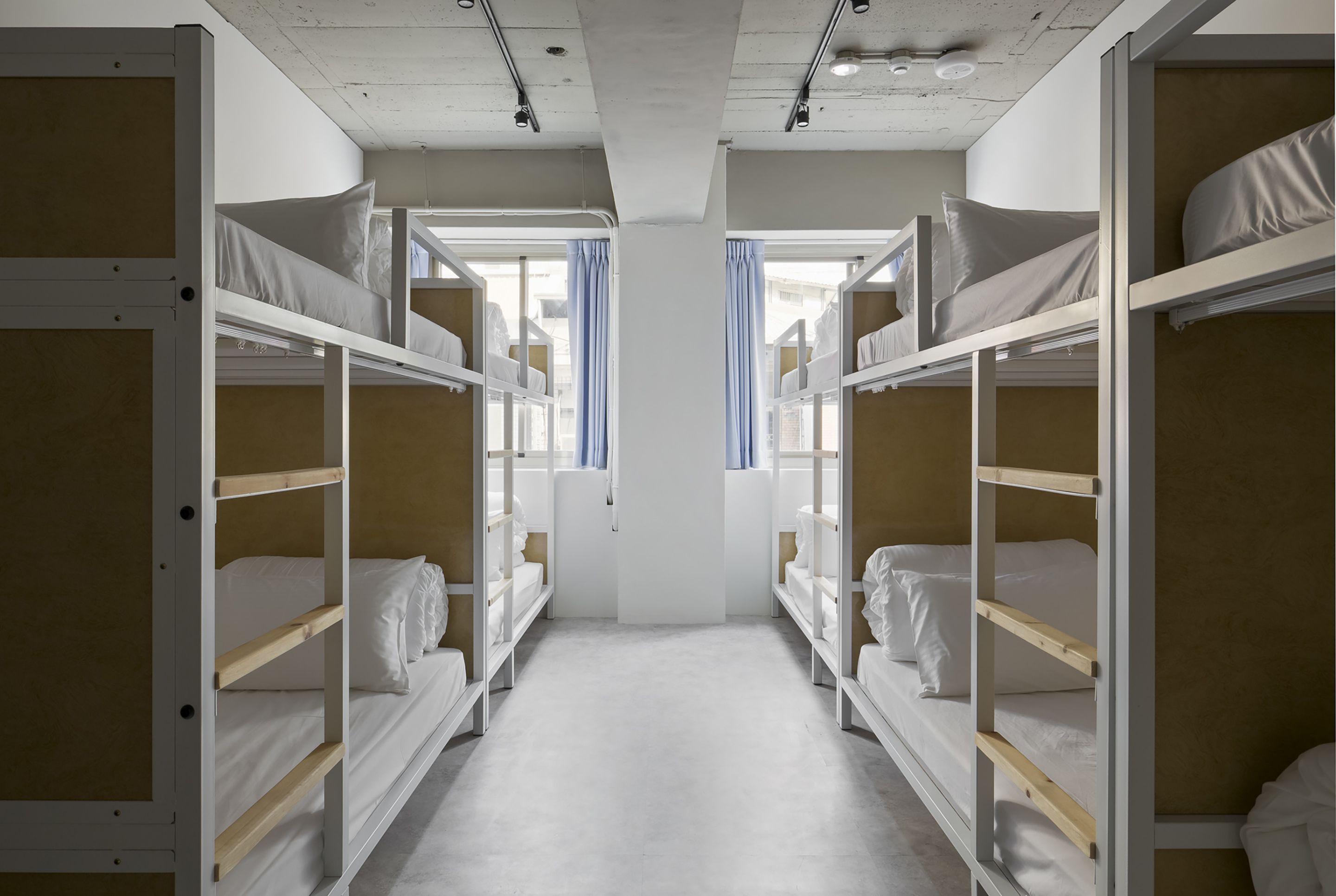 Quarto com beliches, um pilar branco no centro e uma janela com cortinas.
