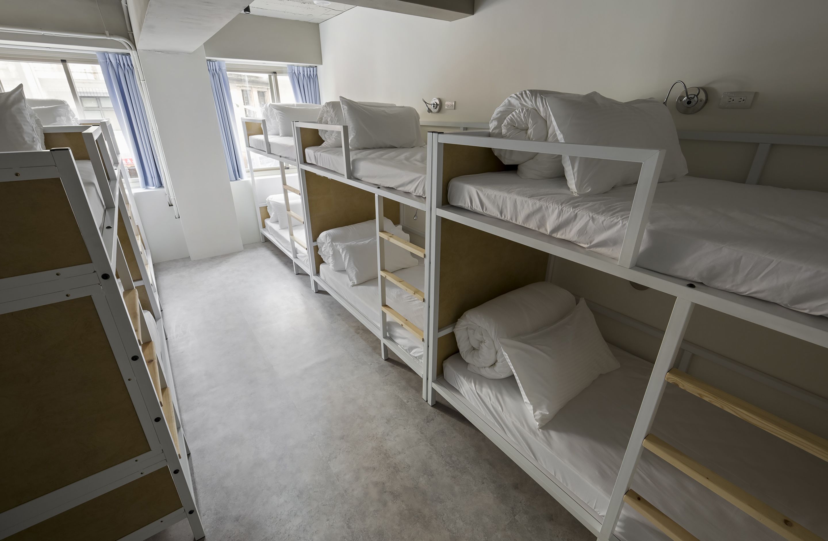 A dormitory room with bunk beds, a window with curtains, and a white wall with a power outlet.