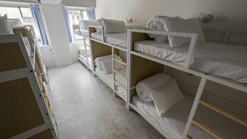 A dormitory room with bunk beds, a window with curtains, and a white wall with a power outlet.