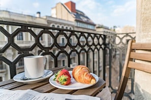 En balkong med ett träbord, en vit kopp, en tallrik med en croissant och en jordgubbe samt en tidning.