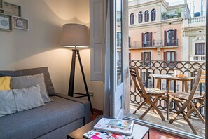 Ein Zimmer mit grauem Sofa, einer Stehlampe, einem Tisch mit Zeitschriften und einem Balkon mit Holzstühlen und einem Tisch.