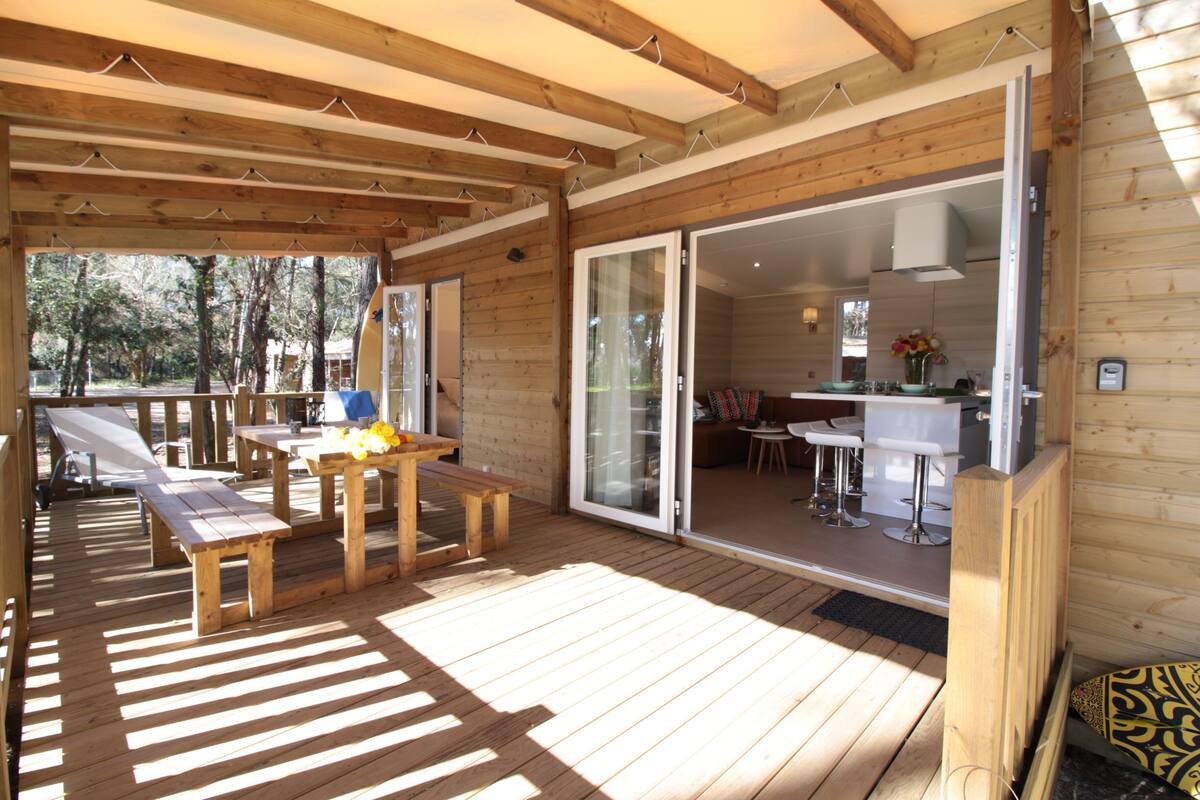 A wooden deck with picnic tables and benches, a glass door leading to a kitchen area with bar stools, and a view of trees outside.