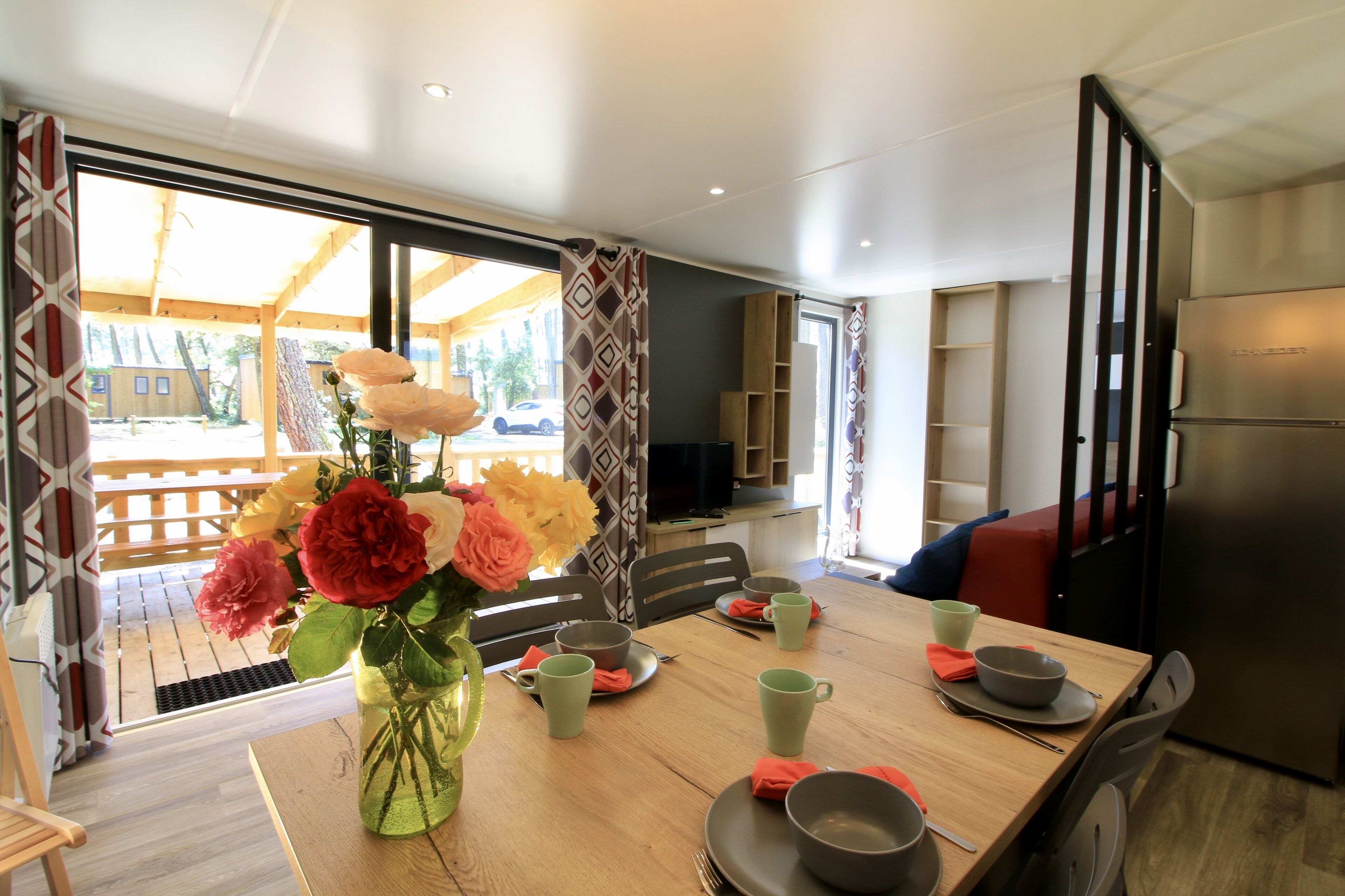 Comedor con mesa de madera, sillas, un florero con flores y cocina moderna con refrigerador.