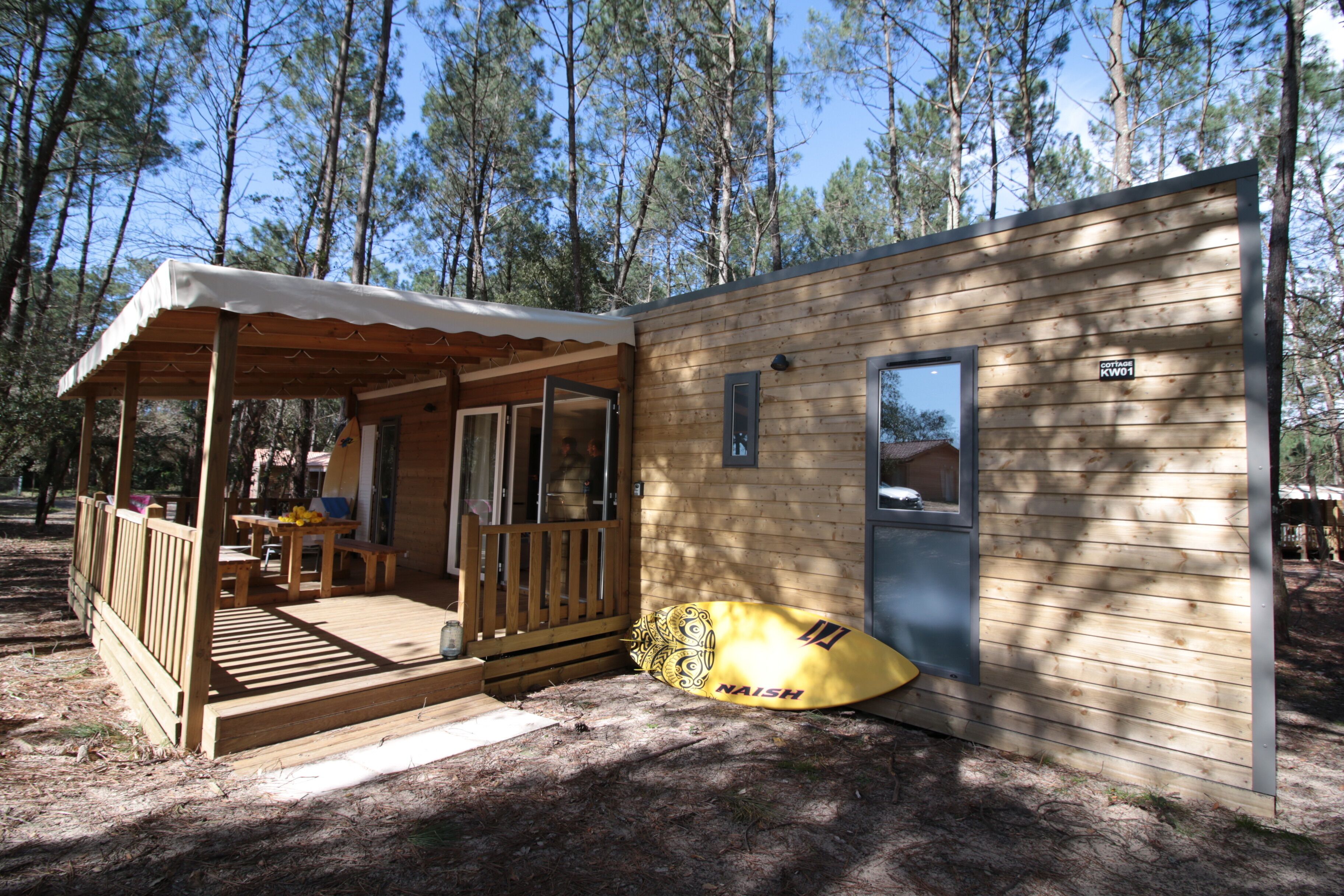 A small wooden cabin with a porch, a dining area, and a kayak labeled "NAISH" by the door.