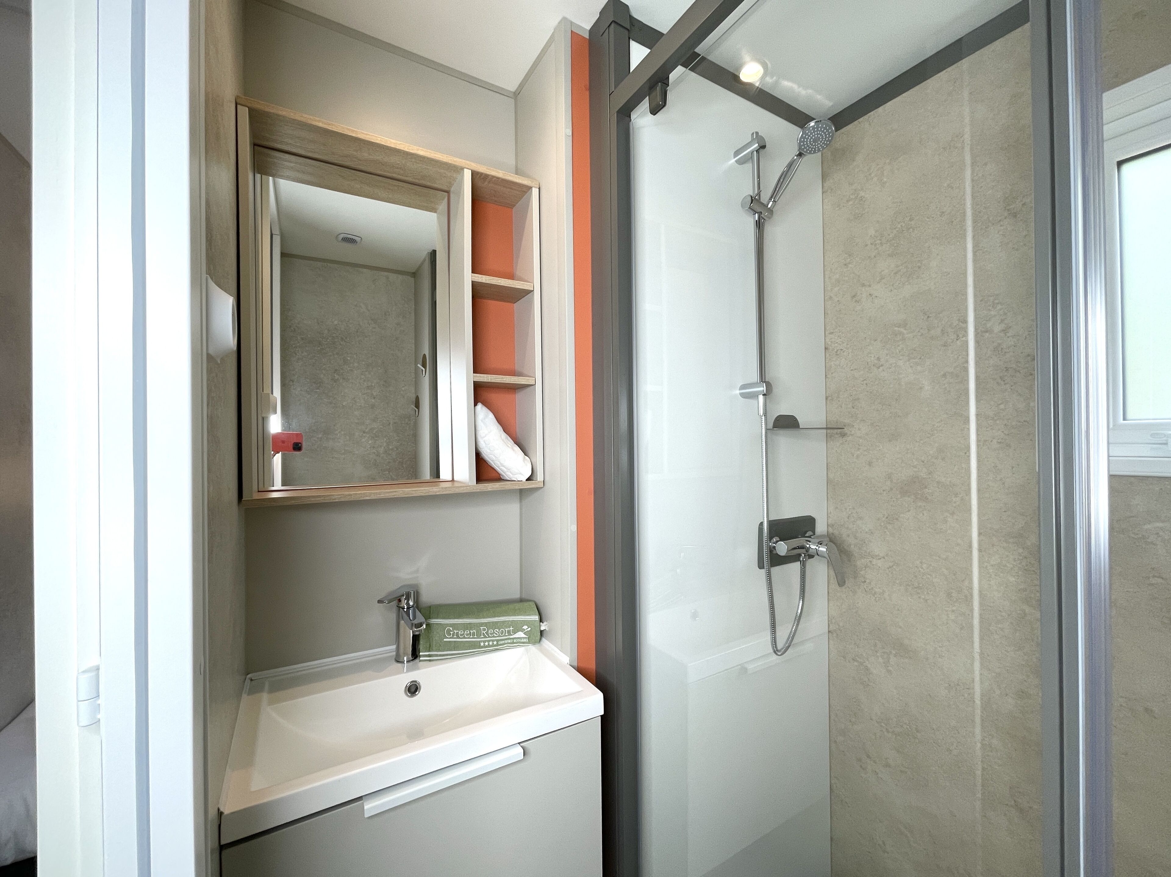 Un baño con ducha de vidrio, lavamanos con dispensador de toallas y espejo con repisas.