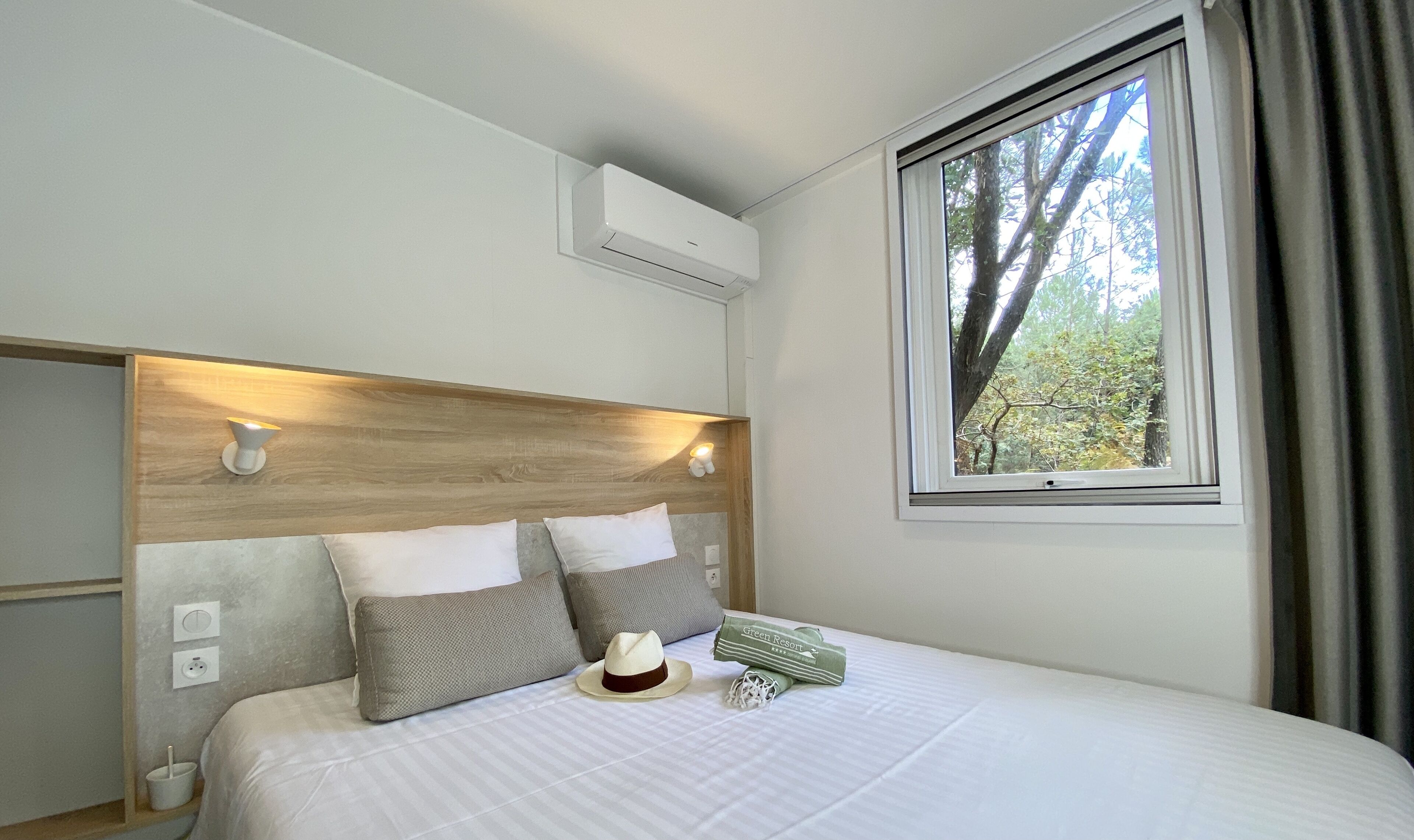 A neatly made bed with a wooden headboard, a window with a view of trees, and a wall-mounted air conditioner.
