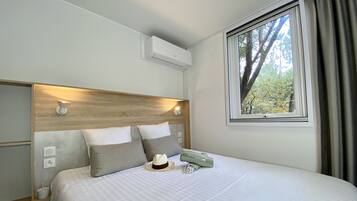 A neatly made bed with a wooden headboard, a window with a view of trees, and a wall-mounted air conditioner.