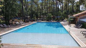 Una piscina rectangular rodeada por una terraza de madera y sillas de descanso, con altos árboles al fondo.