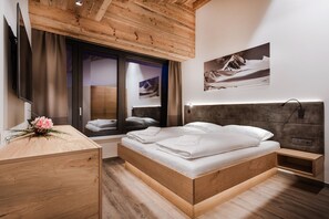 Ein modernes Schlafzimmer mit einem hölzernen Kopfende, einem Bett mit ordentlich zusammengefalteter weißer Bettwäsche, einem Nachttisch aus Holz mit einer rosa Blume, einem Fernseher an der Wand und einem Fenster mit Bergblick.