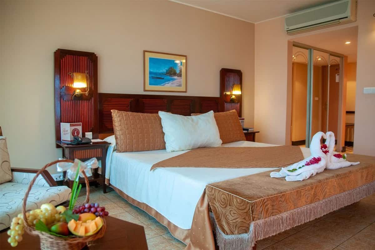 Una habitación de hotel con una cama grande, una mesita de noche con lámpara, un cuadro colgado en la pared y una cesta de frutas.