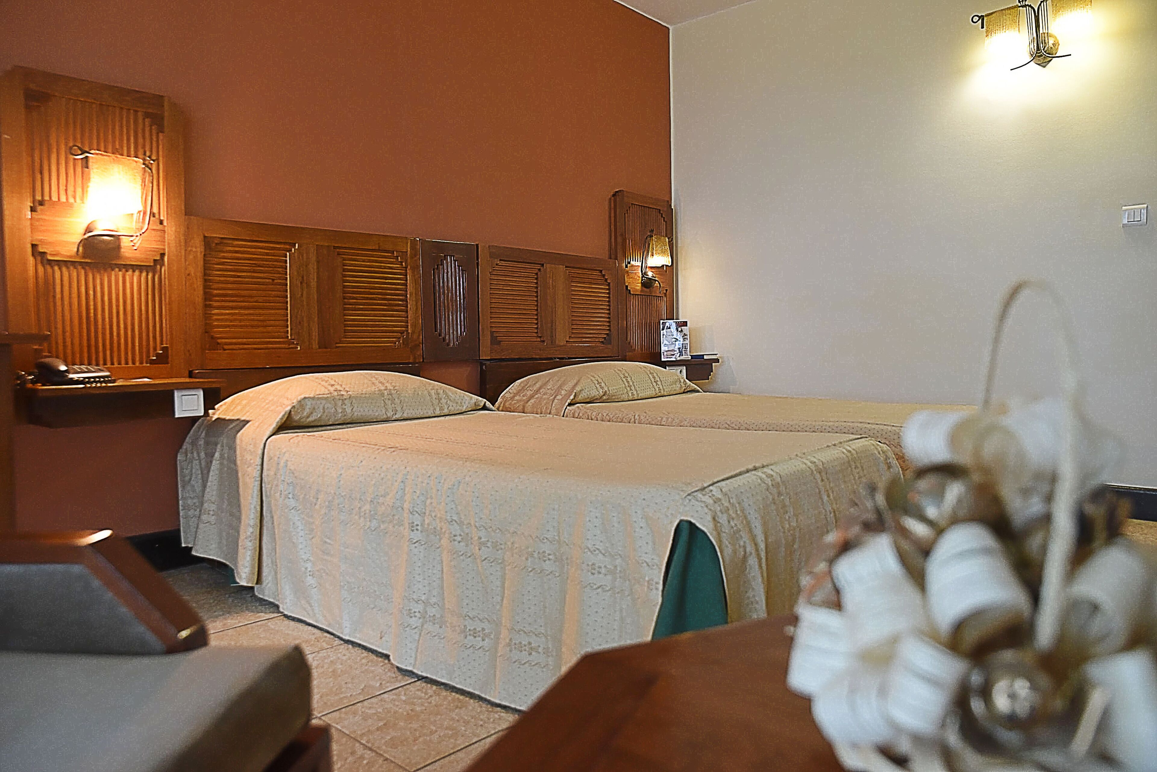 A hotel room with two beds, a wooden headboard, a desk with a telephone, and wall-mounted lamps.