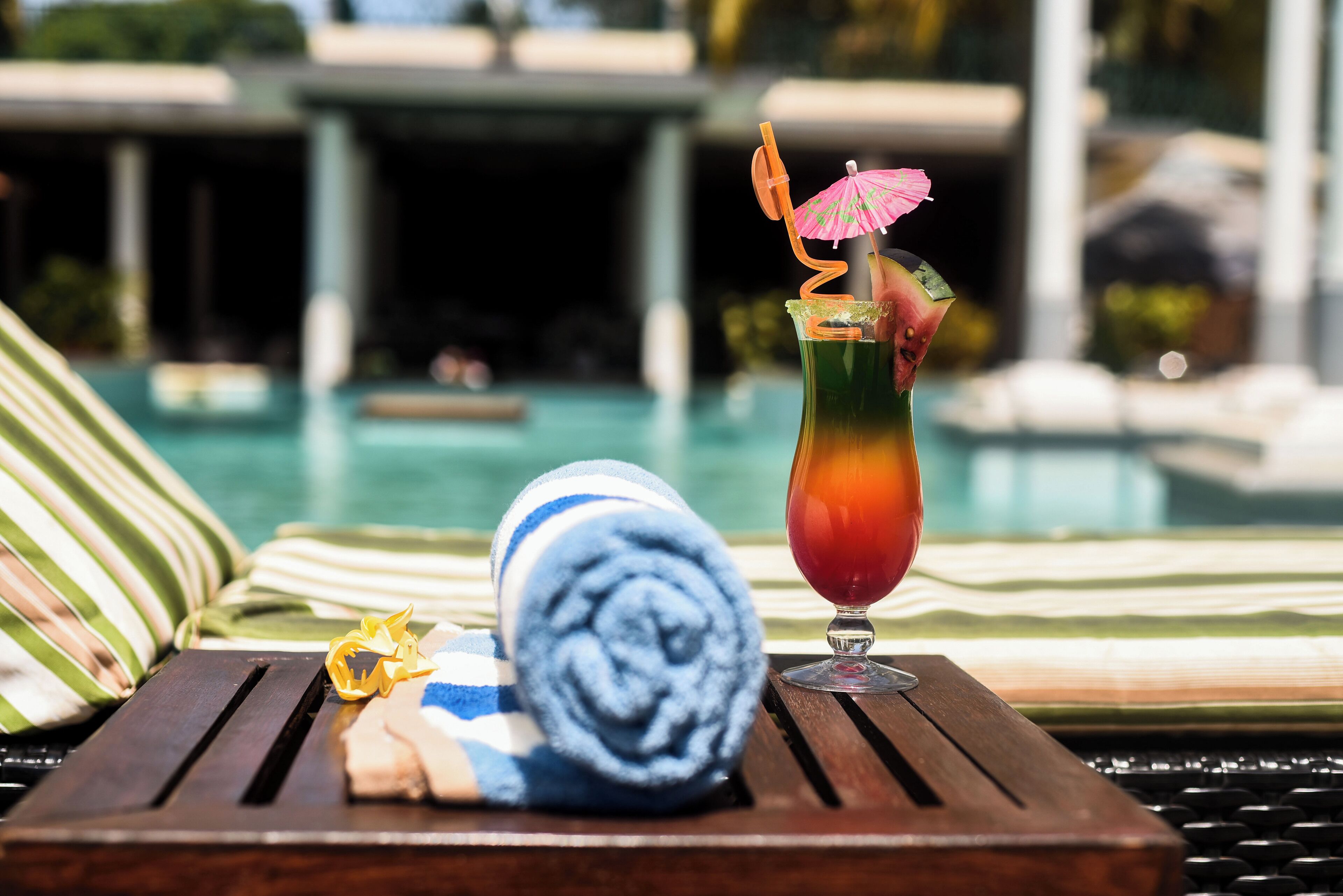 Escena junto a la piscina con una silla de descanso, una toalla enrollada y un cóctel con un parasol decorativo.