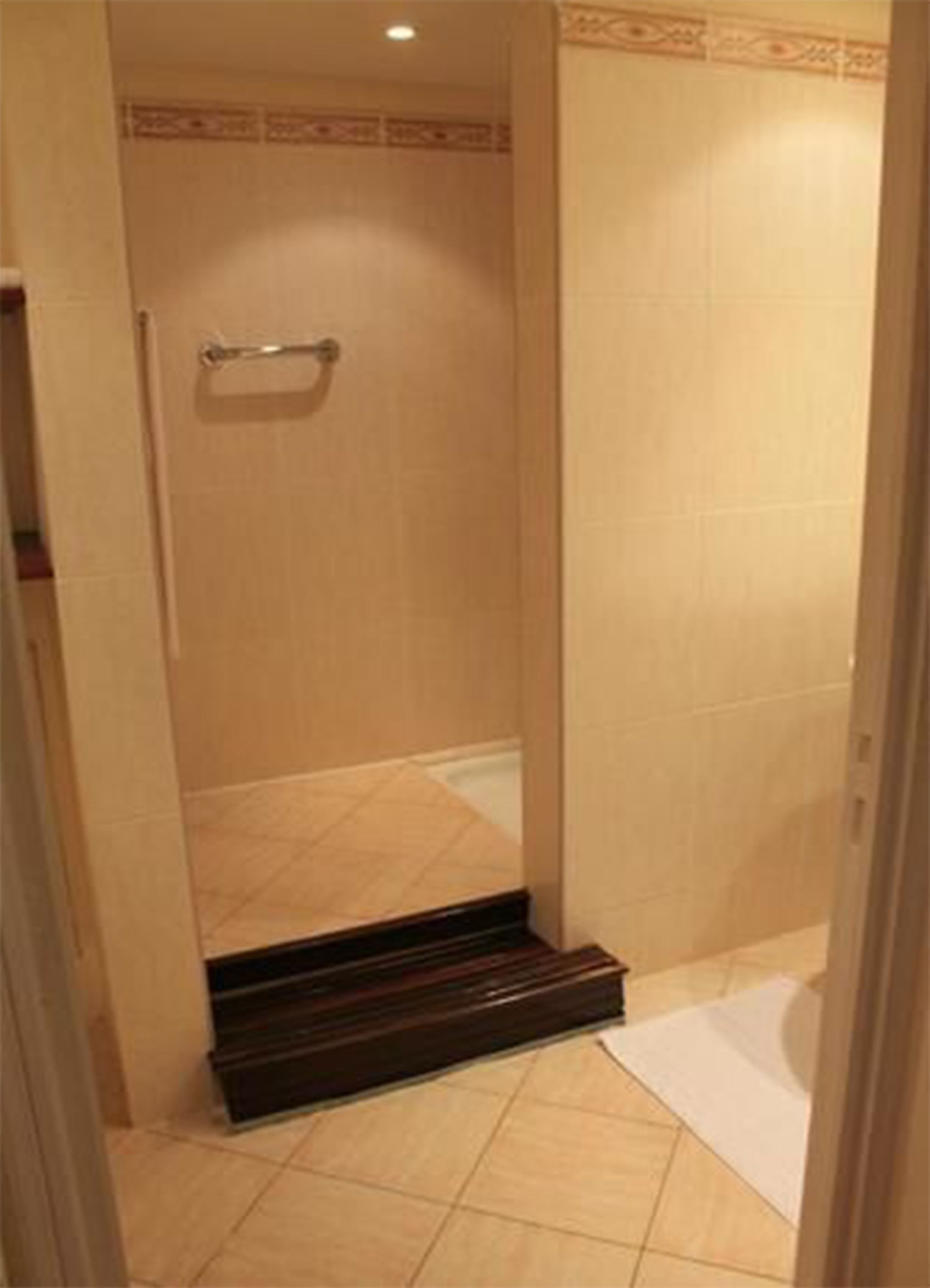 Zona de ducha pequeña y cerrada, con una barra de apoyo de metal, paredes de azulejos y un escalón de madera para entrar.