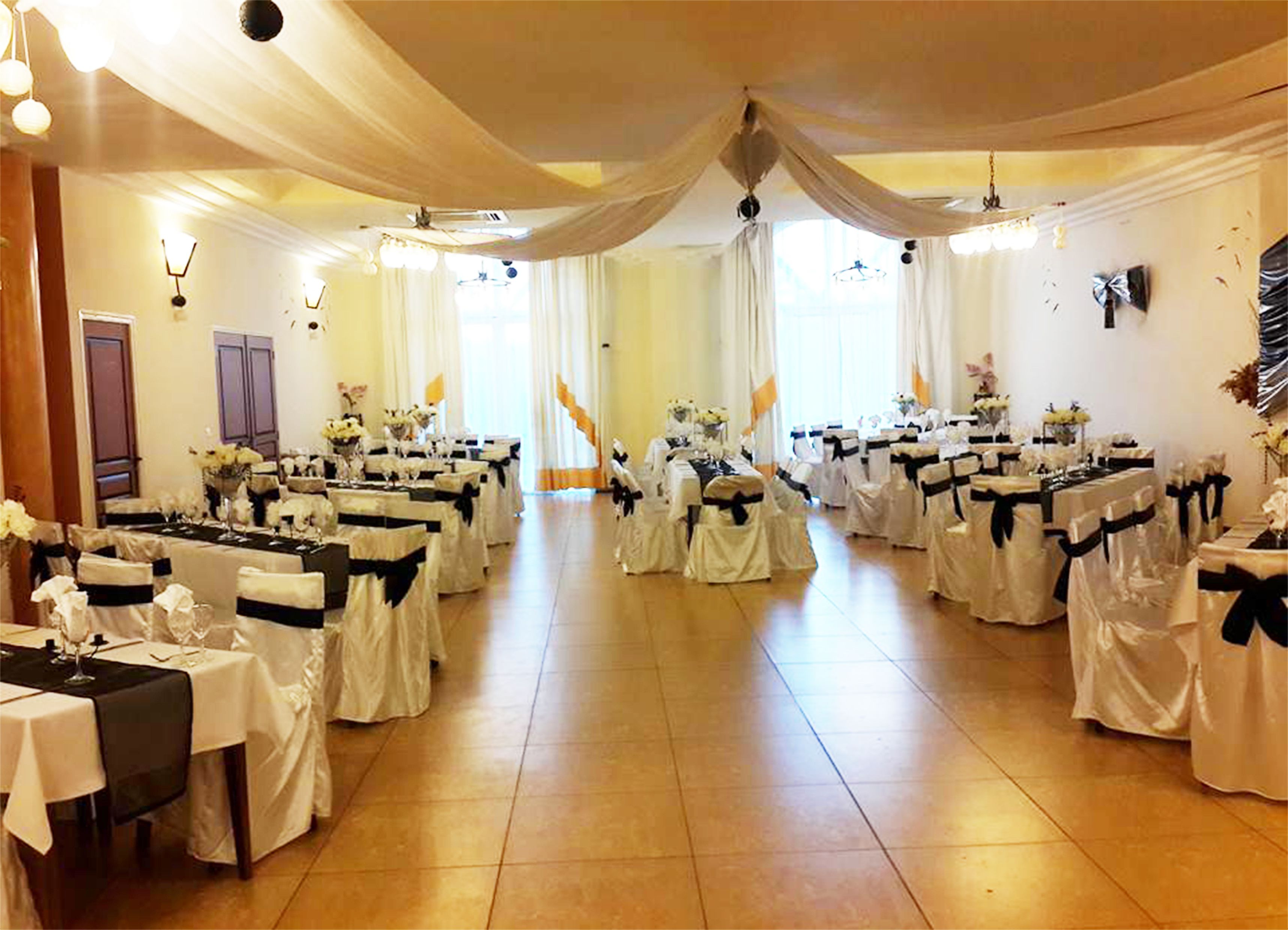 Uma sala de banquetes com mesas montadas para um evento formal, cobertas com toalhas brancas e fitas pretas, e decorada com arranjos florais.