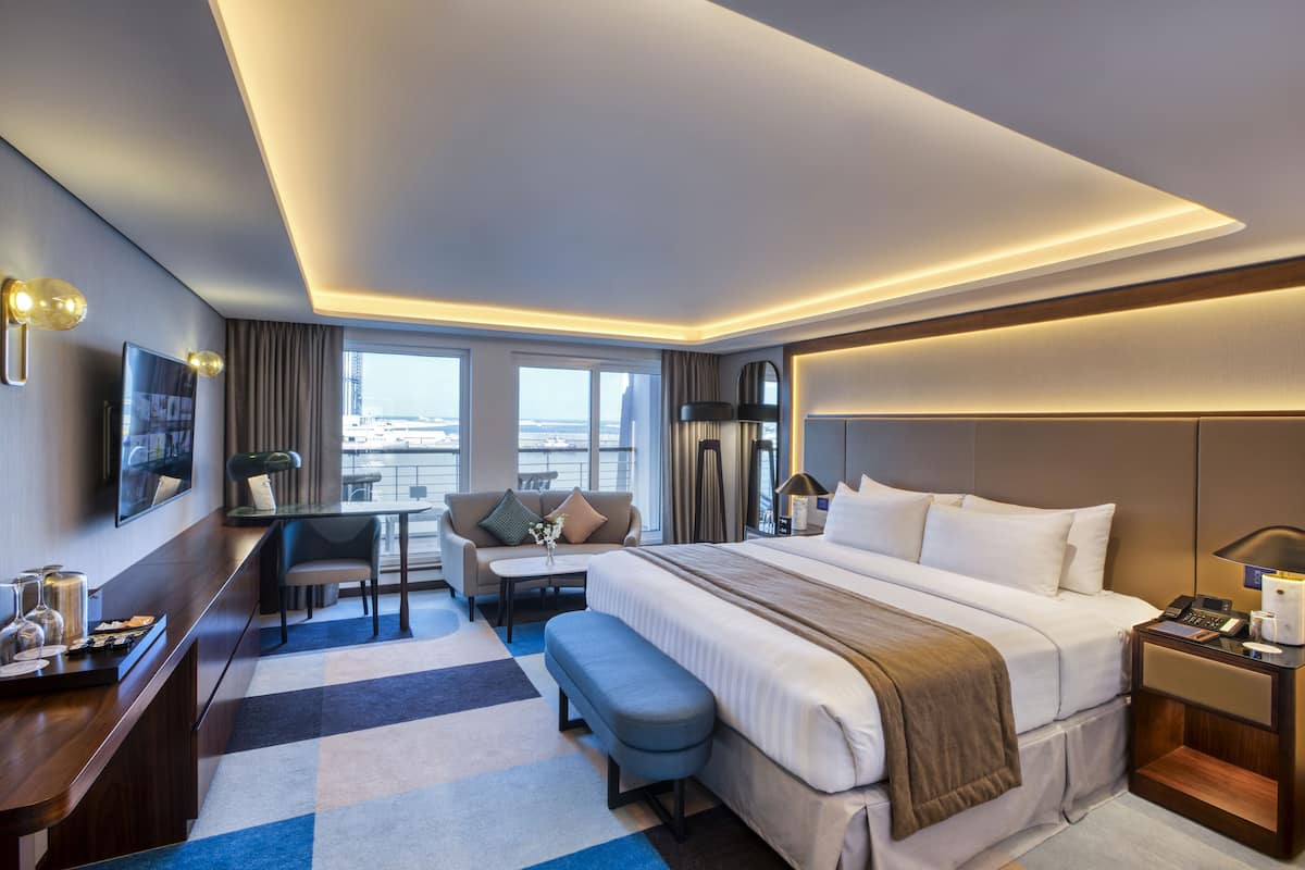 A modern hotel room with a large bed, a desk, a sofa, and a view of the city.
