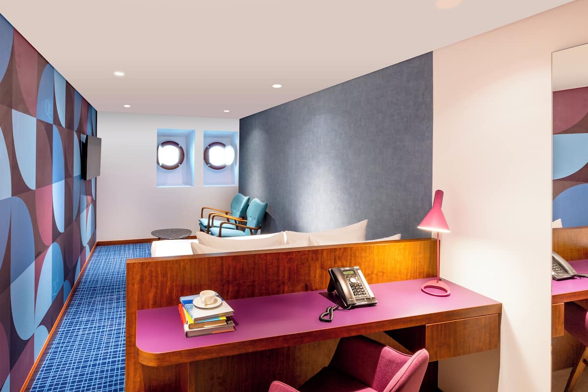 A modern hotel room with a wooden desk, a bed with blue and white bedding, a small table with a lamp, and a wall with geometric patterns.