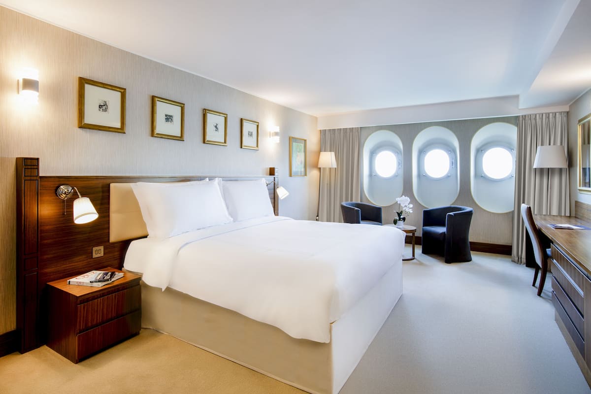 A hotel room with a large bed, bedside tables, a desk, and a chair. There are framed pictures on the wall and a window with curtains.