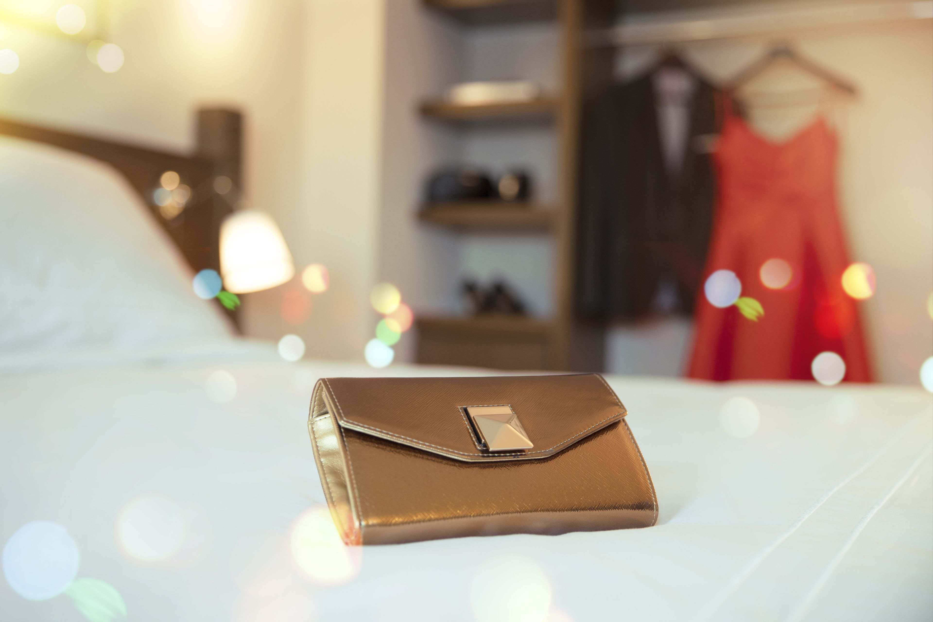 Uma clutch marrom bem dobrada sobre uma cama, com um fundo desfocado mostrando um vestido vermelho e prateleiras.