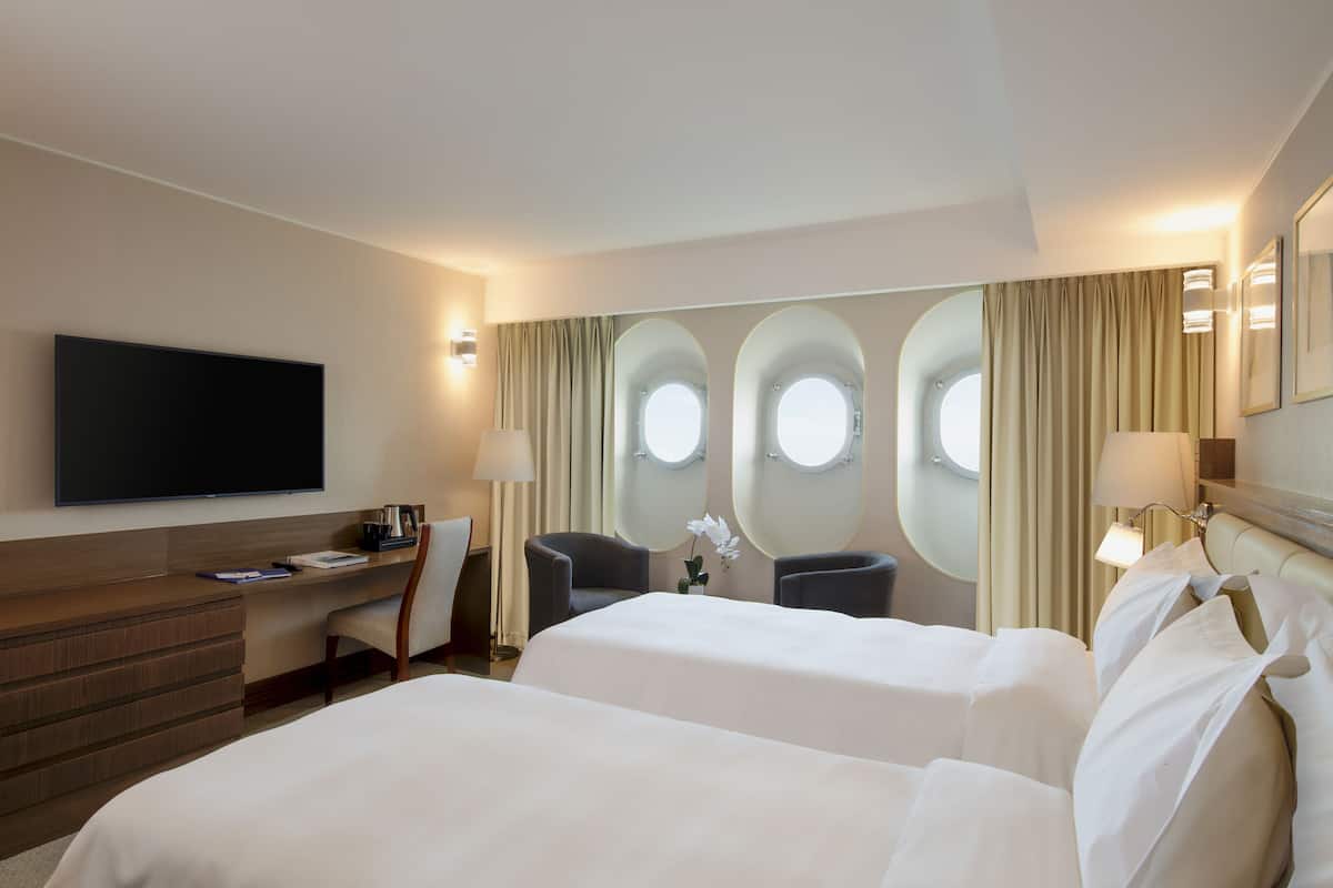 A hotel room with two beds, a desk, a TV, and a window with curtains.