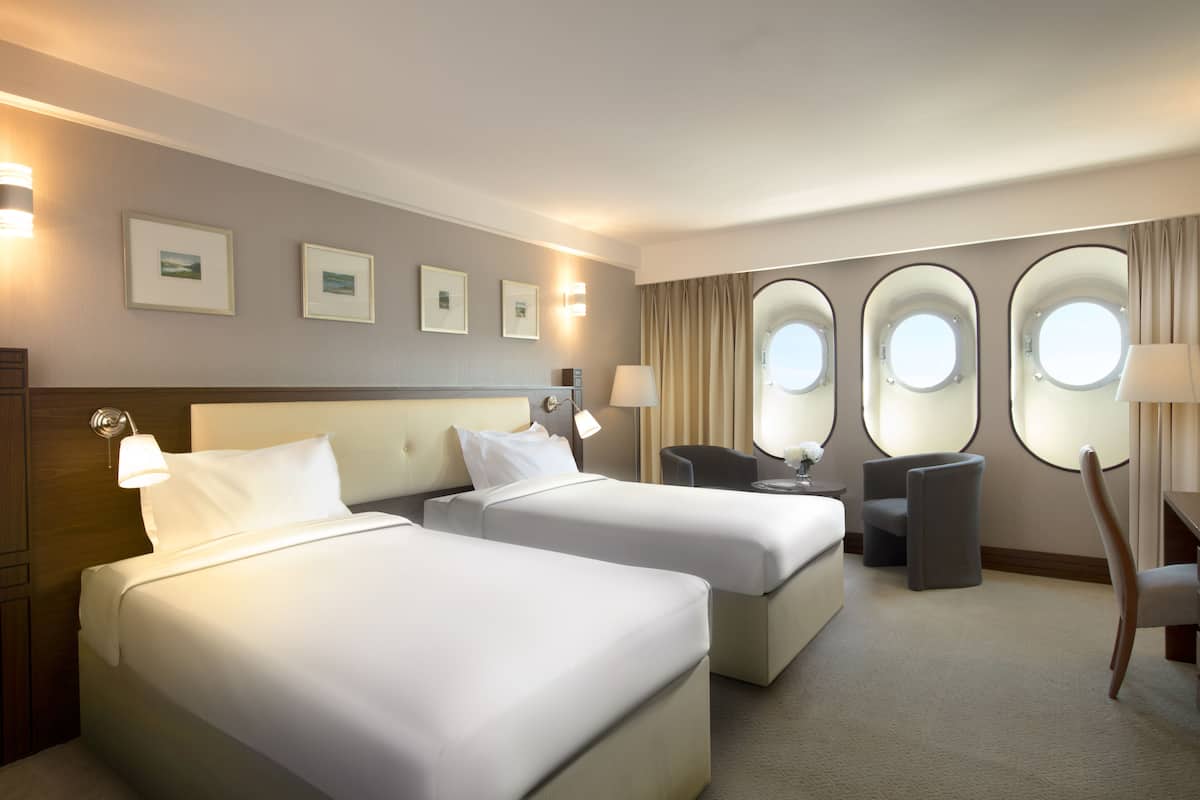 A hotel room with two beds, a desk, a chair, and a window with a view.