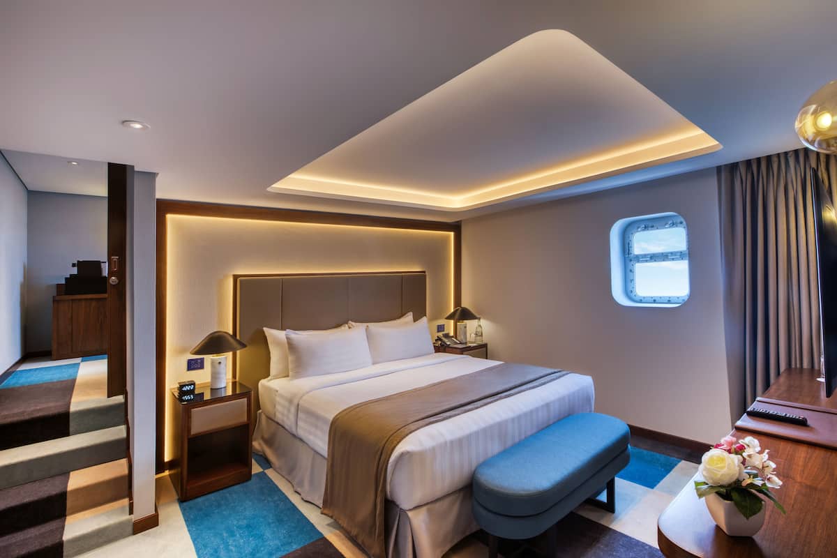 A modern hotel room with a large bed, a desk, a chair, a small table, a nightstand, a window, and a staircase.