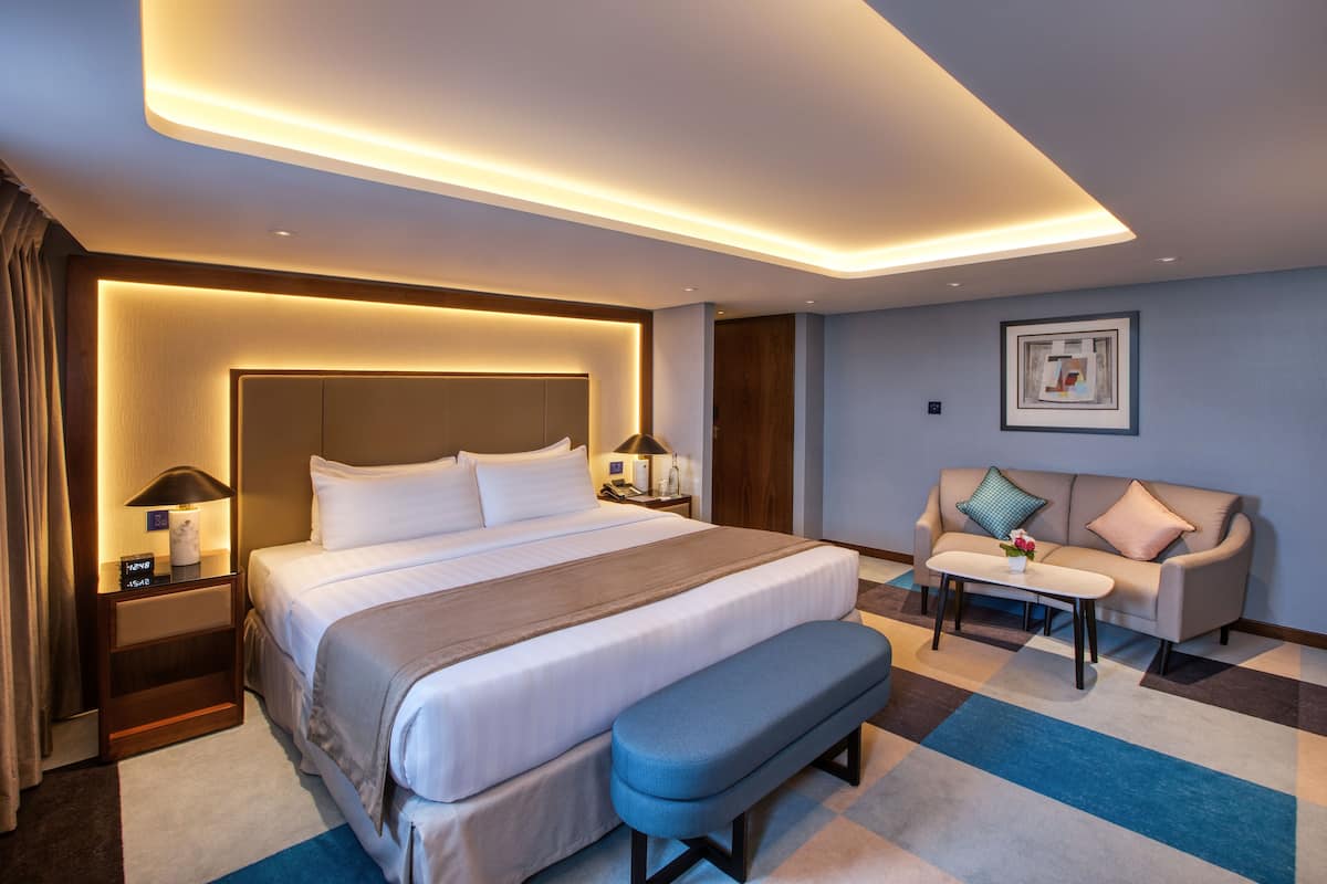 A modern hotel room with a large bed, bedside tables, a bench, a sofa, and a small table.