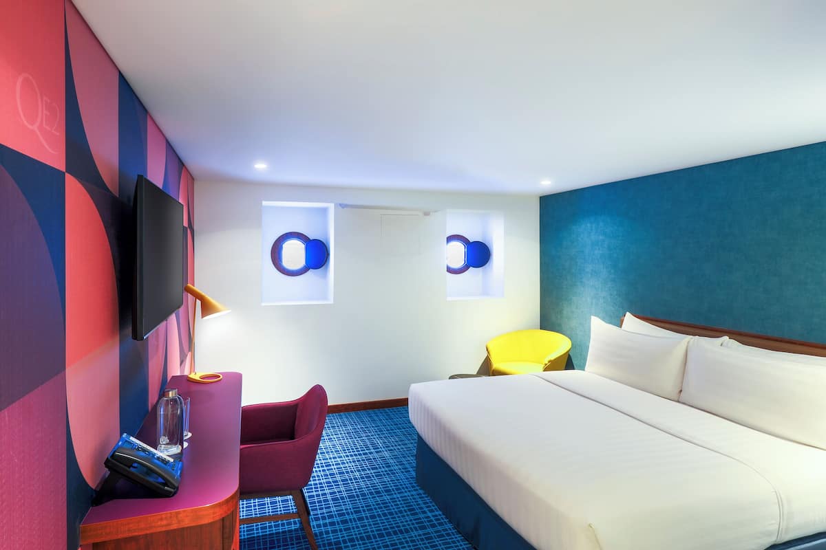 A hotel room with a bed, a desk with a chair, a TV, and two wall-mounted circular windows.
