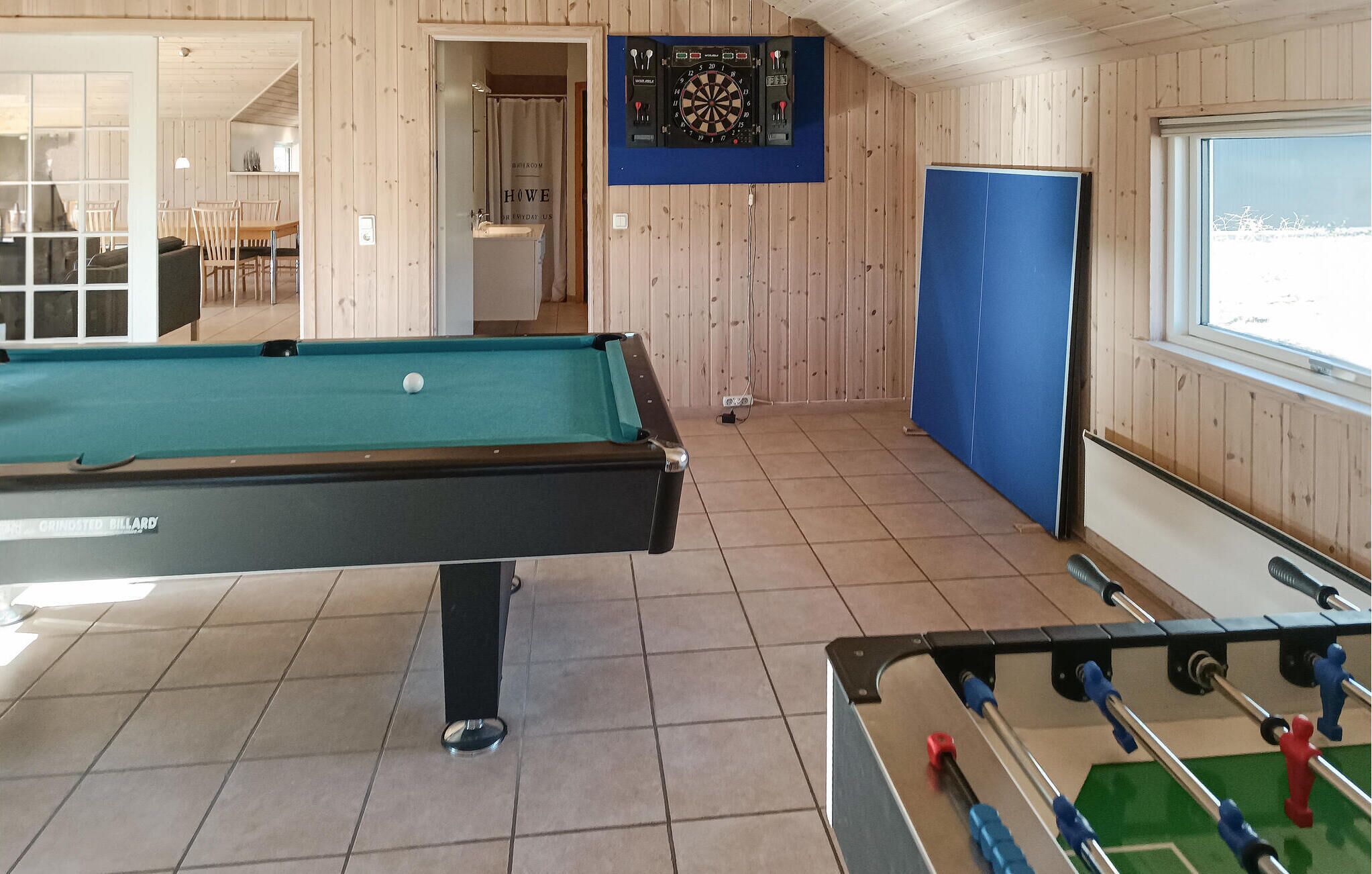 A pool table with a dartboard and foosball table in a room with wooden walls and a large window.