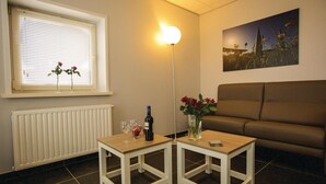 A living room with a brown sofa, two white tables with drinks and a vase of flowers, a radiator, a window with blinds, and a floor lamp. - Lovely home in Retranchement with WiFi (Retranchement)