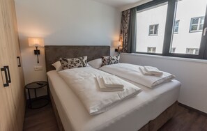 A double bed with white linens and pillows, a bedside table with a lamp, and a window with curtains.