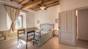 Ein Schlafzimmer mit Steinwand, Holzboden, einem Bett mit Metallgestell, einem Holzstuhl, zwei Nachttischen mit Lampen und einer Tür.