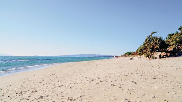 Ein weiter Sandstrand mit Fußspuren, einem klaren blauen Himmel und einer entfernten Brücke.