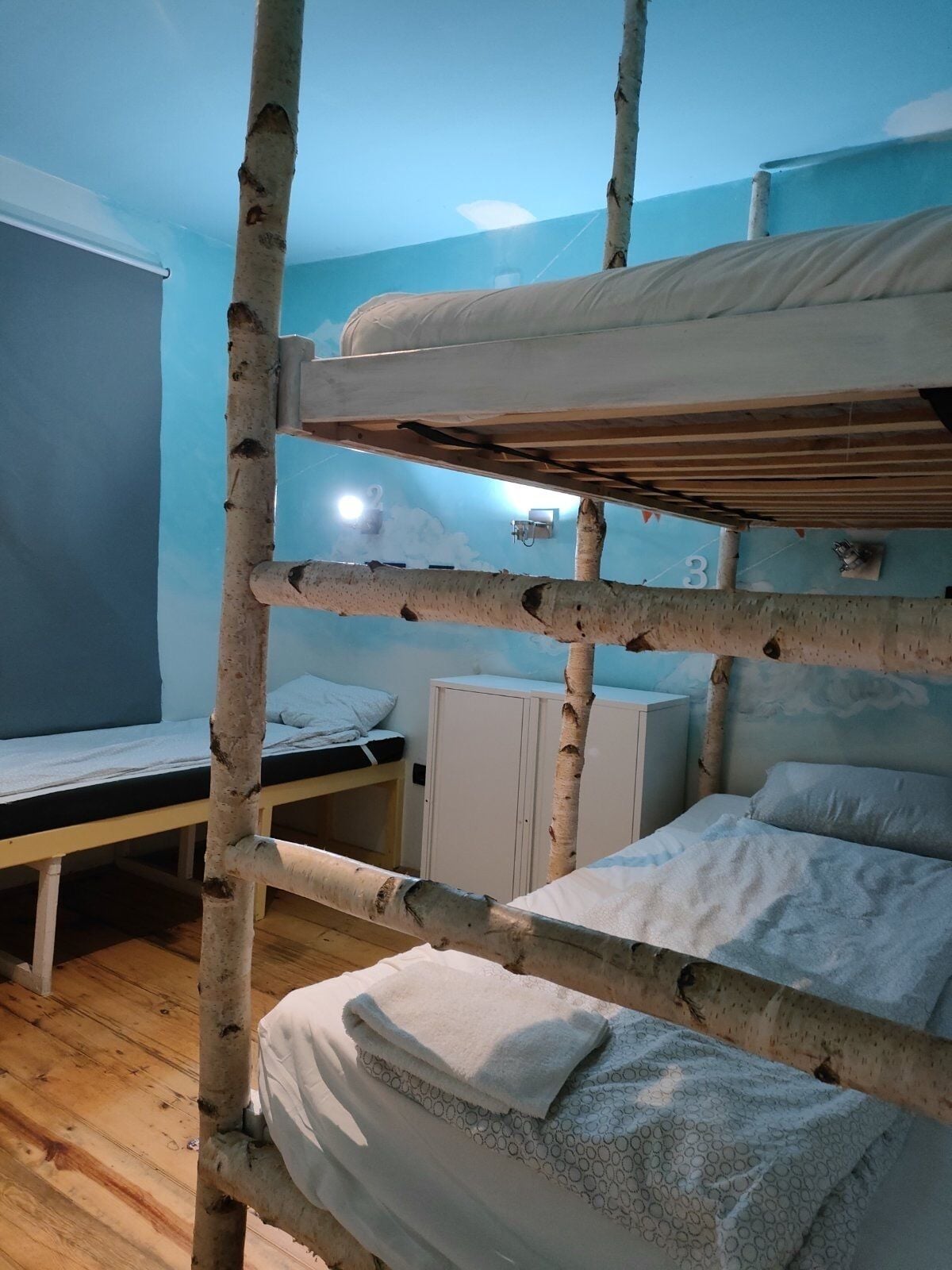 A bunk bed with a wooden frame, a single bed with a white mattress, and a cabinet in a room with blue walls and wooden flooring.