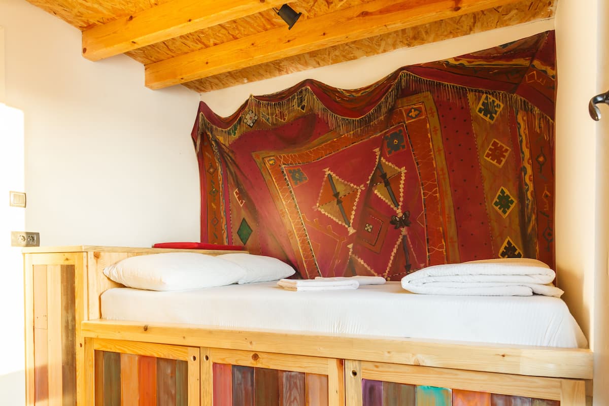 A wooden bed with a white mattress and pillows, a red patterned tapestry, and a wooden ceiling.