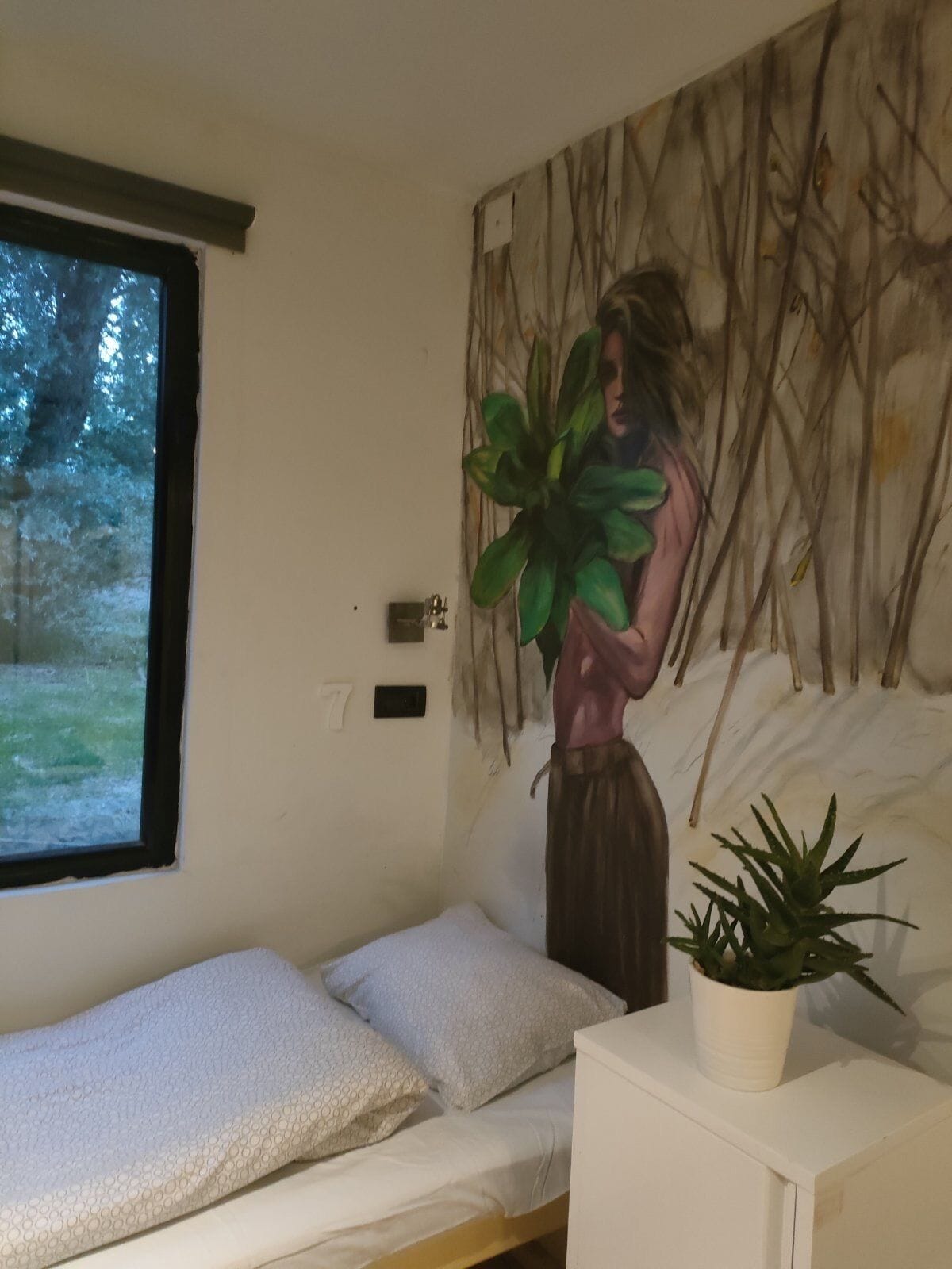 Un dormitorio con una cama, una ventana, un interruptor de luz en la pared y una planta en maceta.