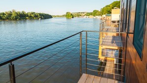 A river view from a wooden deck with a metal railing, overlooking a calm river and a distant shoreline with buildings and trees. - ArkaBarka Floating Hostel and Apartments (Belgrade)