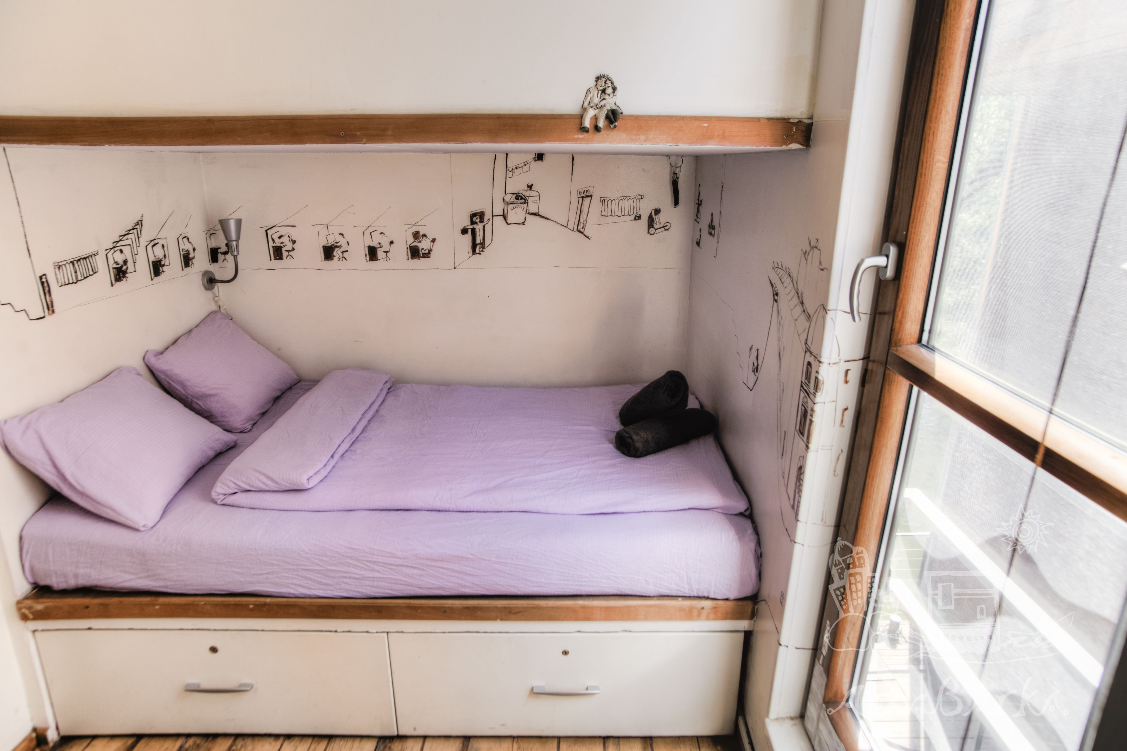 A small, single-bed room with a wooden headboard, a white wall with sketches, and a window with a view.