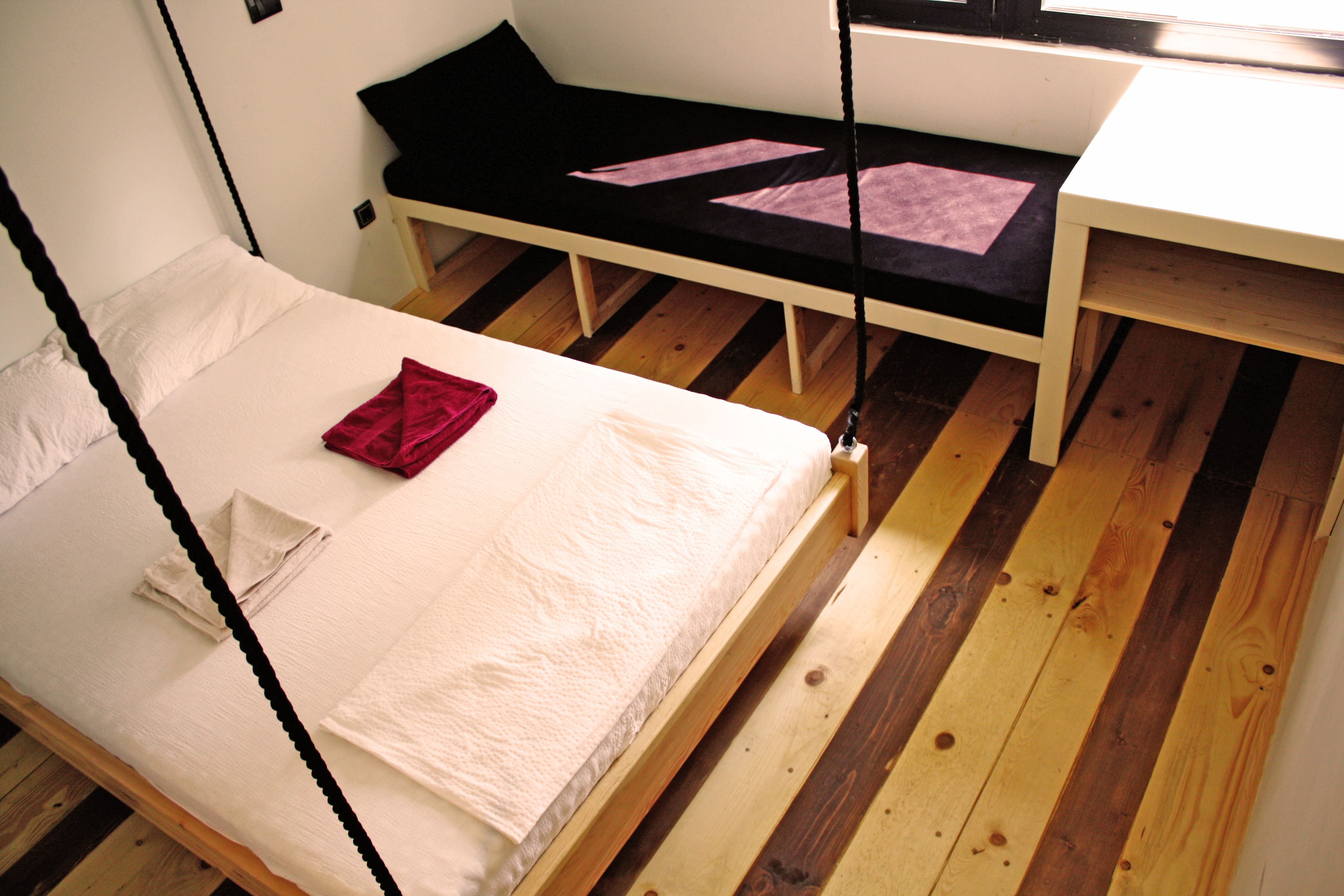 A single bed with a white mattress and a red blanket, a small table with a black surface, and a black bench with pink cushions.