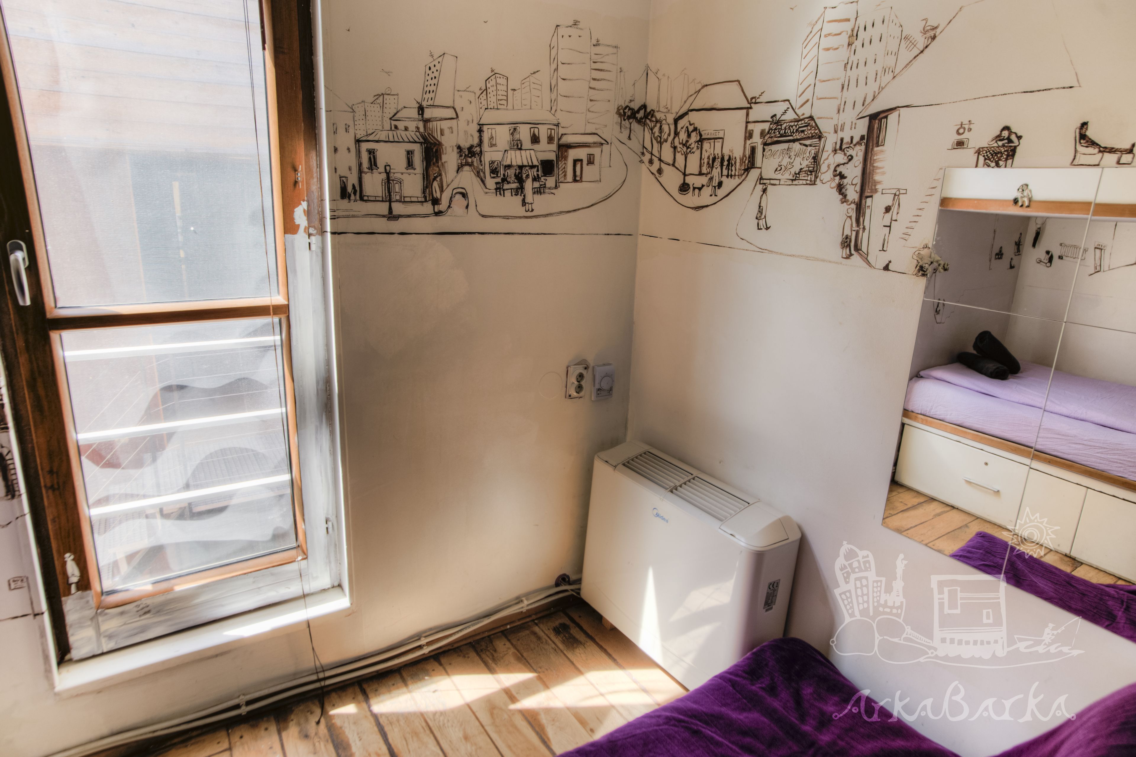 Una habitación con ventana, un radiador, una cama con manta púrpura y una pared con un dibujo de un paisaje urbano.