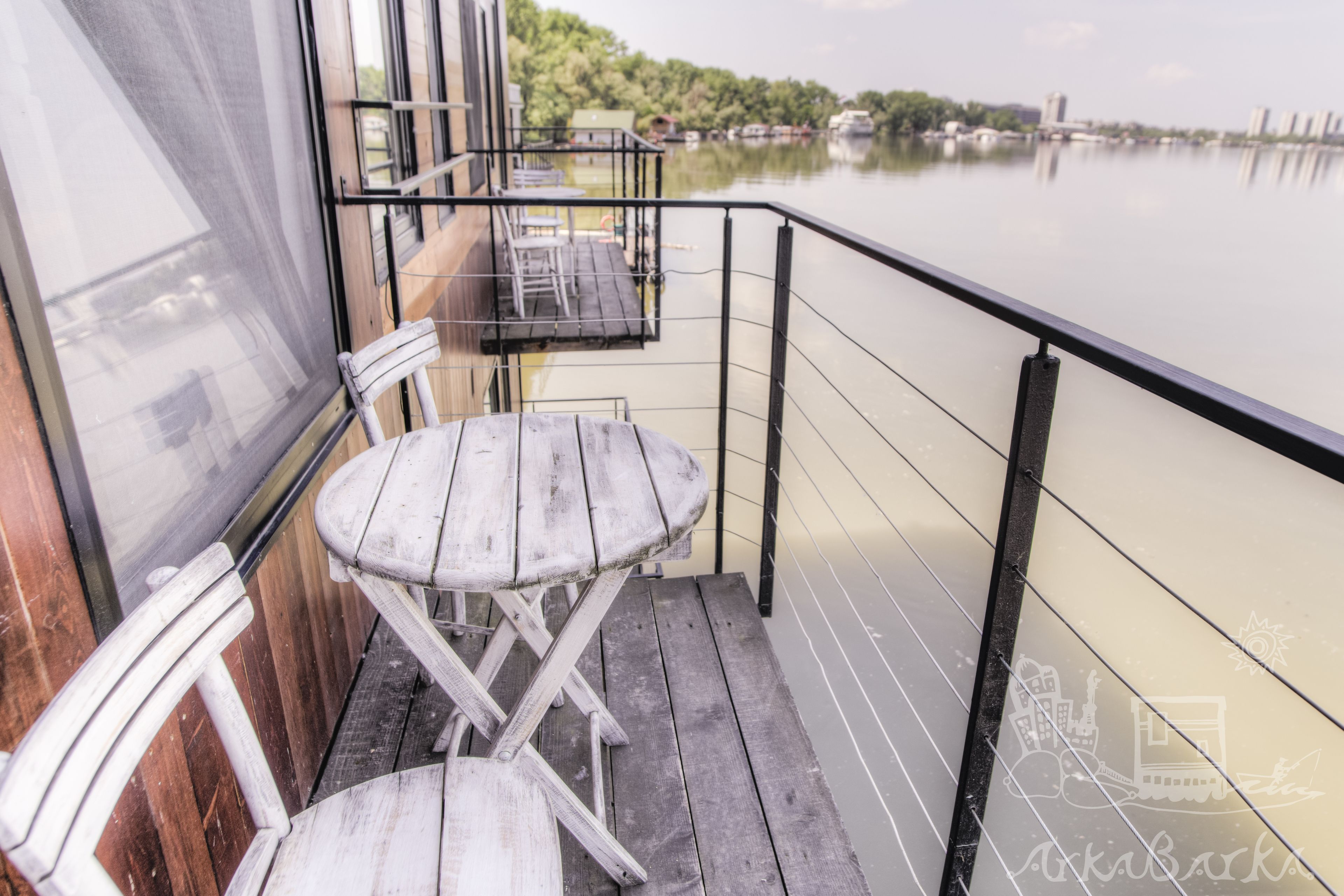 Une terrasse en bois avec une table et des chaises, donnant sur une étendue d’eau calme, avec des bâtiments visibles au loin.