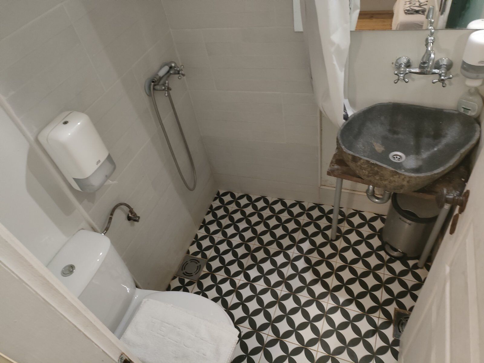 Et lille badeværelse med toilet, håndvask og bruser. Gulvet er belagt med et geometrisk sort-hvidt mønster.