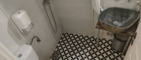 Et lille badeværelse med toilet, håndvask og bruser. Gulvet er belagt med et geometrisk sort-hvidt mønster.