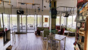 A spacious indoor area with wooden flooring, tables, chairs, and a large window offering a view of the outdoors. - ArkaBarka Floating Hostel and Apartments (Belgrade)