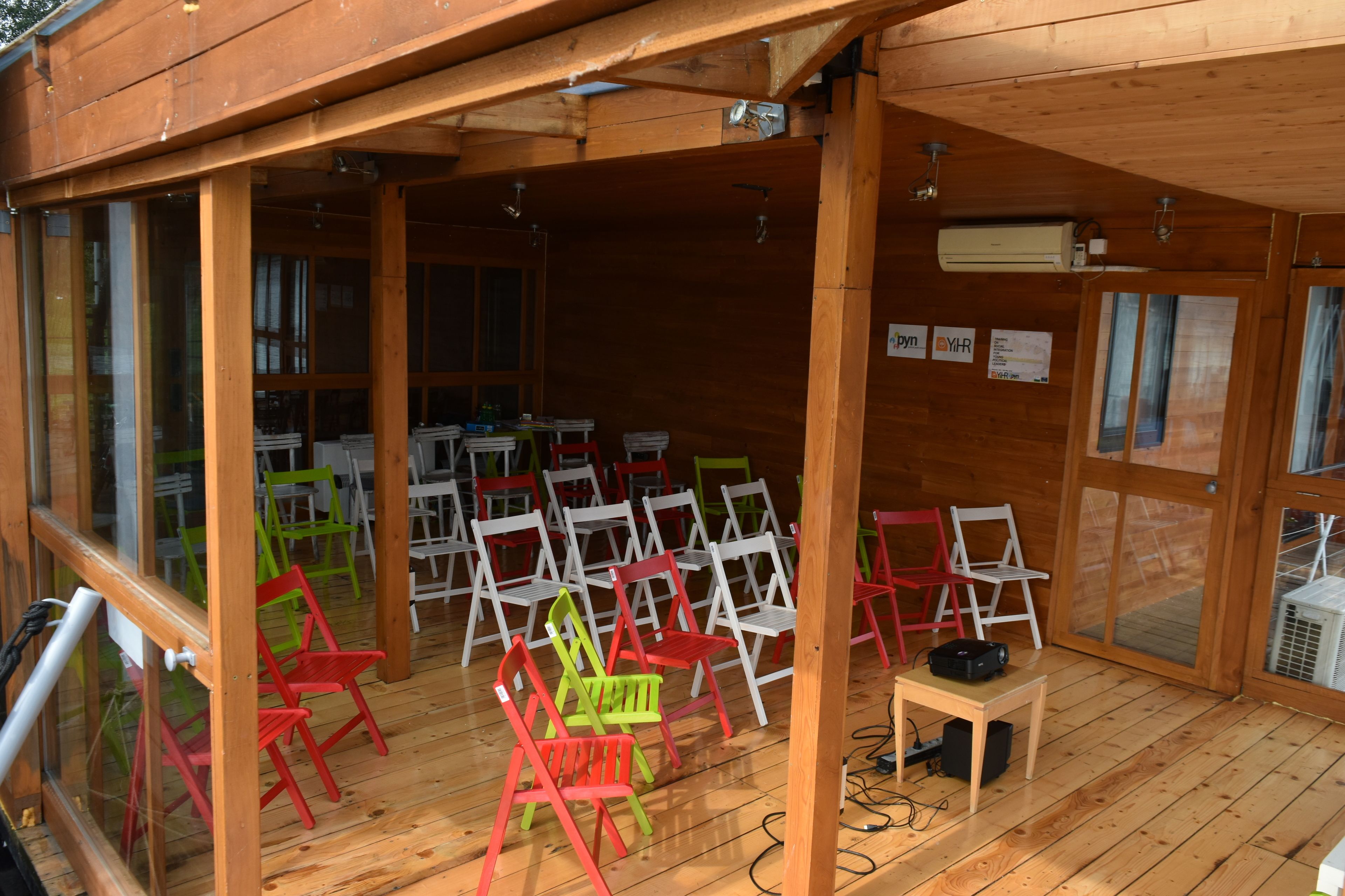 A covered outdoor seating area with multiple wooden chairs and tables, an air conditioning unit, and a wooden ceiling structure.
